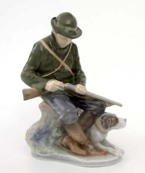 Auf ovalem Sockel sitzt auf einem Baumstumpf der Jäger seine Flinte ladend zu seinen Füßen der Jagdhund. Manufakturmarke, Modell-Nr.
