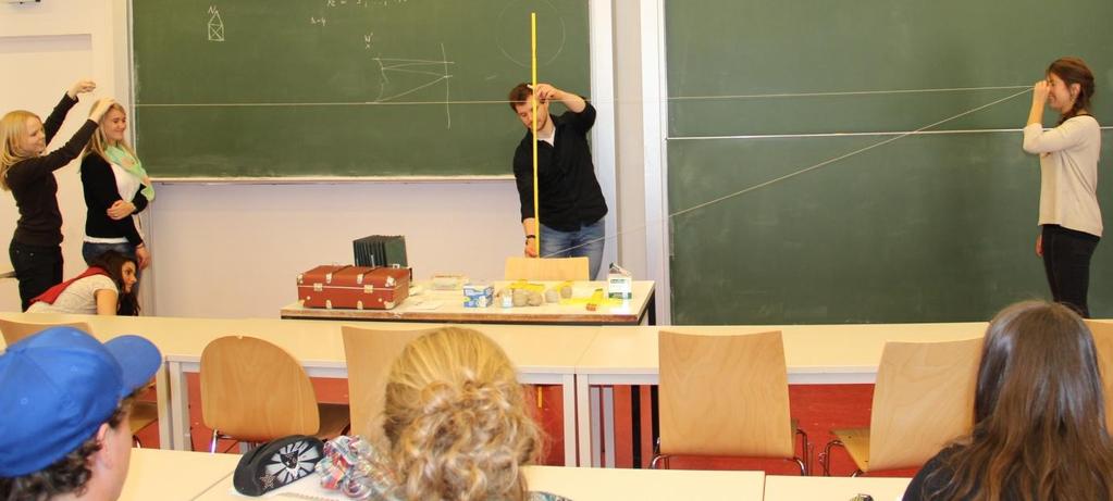 Ein Schüler hält einen Meterstab, der den Spiegel repräsentiert. Zwei weitere Schüler stellen sich mit dem Blick zueinander in gleicher Entfernung von dem Meterstab auf.