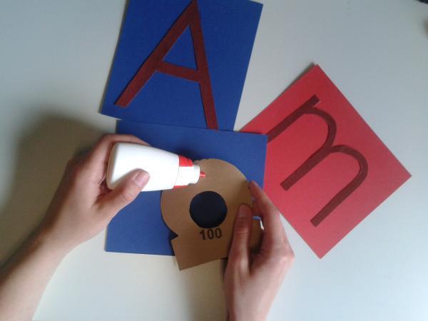 Die Buchstabenformen aus dem Sandpapier ausschneiden und auf den Karton kleben. Hierbei auf die Farben achten: Vokale auf blauen Karton, Konsonanten auf roten Karton.
