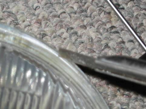Schabe/kratze jetzt vorsichtig mit dem Kleinen Schraubendreher die graue Klebe- und Dichtungsmasse am Scheinwerferrand ab (Muss