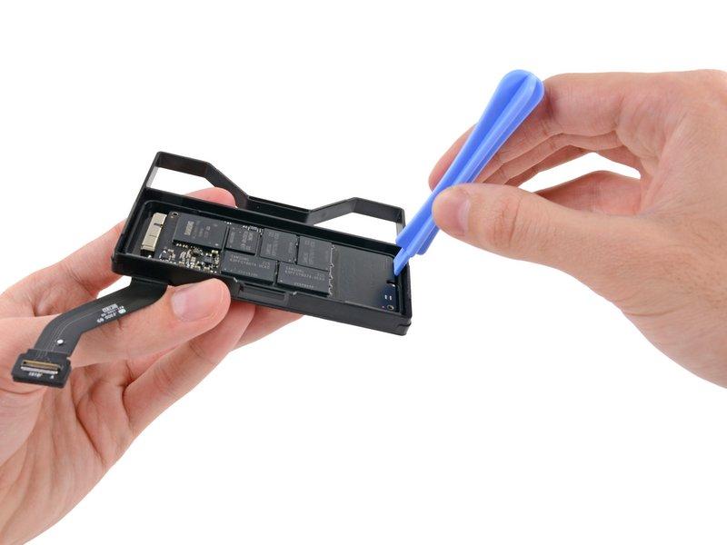 Schritt 14 Setze die Kante eines Plastiköffnungswerkzeugs zwischen SSD und SSD Einschub ein, und zwar auf der Seite gegenüber vom Sockel des SSD Einschubs.