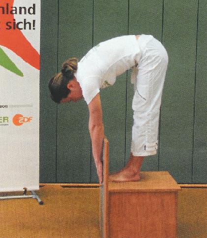 Stand and Reach Die Testperson steht ohne Schuhe auf einer Bank, an dem von der Standfläche aus nach oben und unten eine Messskala angebracht ist.