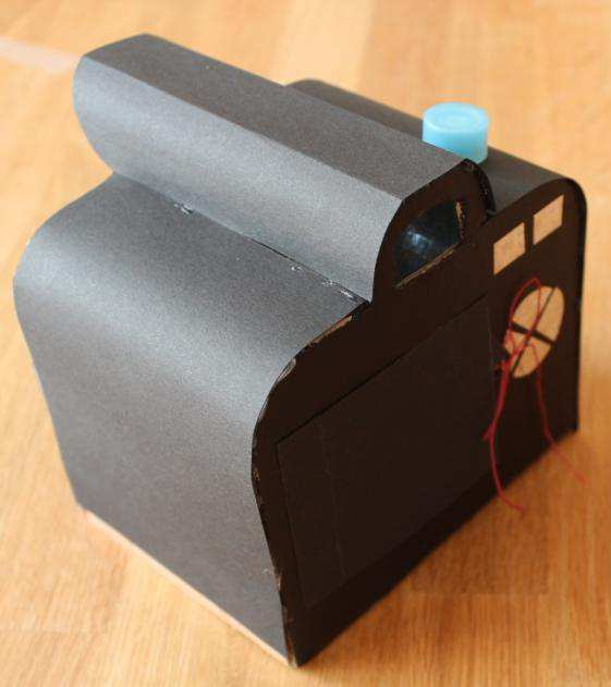 Die Rückseite der Kamera mit der schwarz bemalten Seite nach unten auf eine Unterlage legen. Die kleine Schachtel mit der Heißklebepistole an der unteren Kante aufkleben.