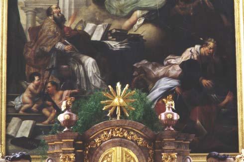 Augustinus, der im bischöflichen Messornat erhöht an einem Schreibpult mit zahlreichen Büchern sitzt. In der rechten Hand hält er eine Schreibfeder, in der linken ein brennendes Herz.