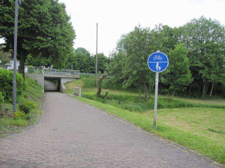 Am Bach dahinter spielen die Kinder gerne. Wenn hier ein kleiner Steg wäre, könnte man die Füße ins Wasser halten. Dass das Schild beklebt ist, fanden die Kinder nicht gut.